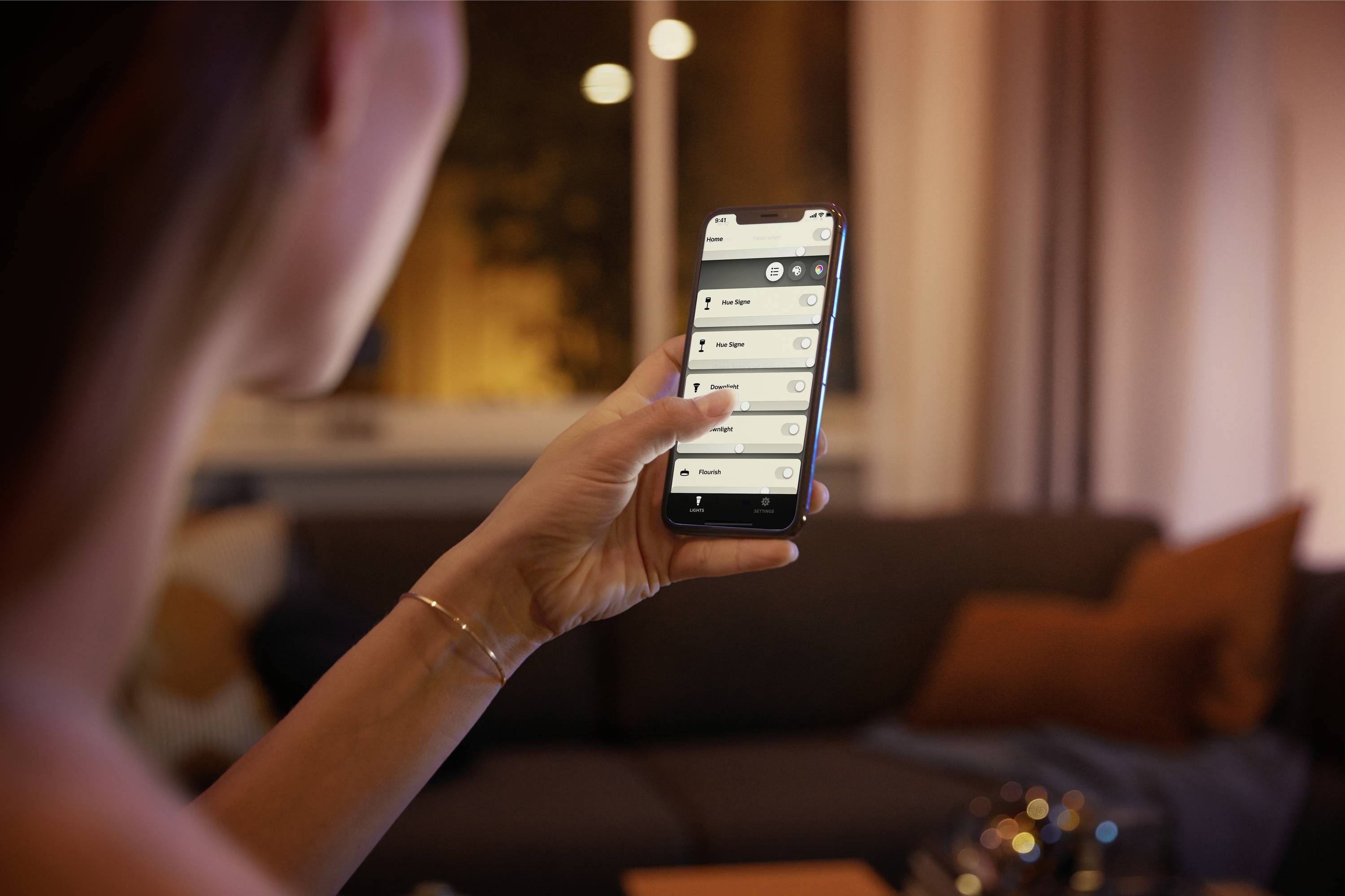 A person is holding a smartphone with a smart home device control app. A cosy living room is visible in the background.