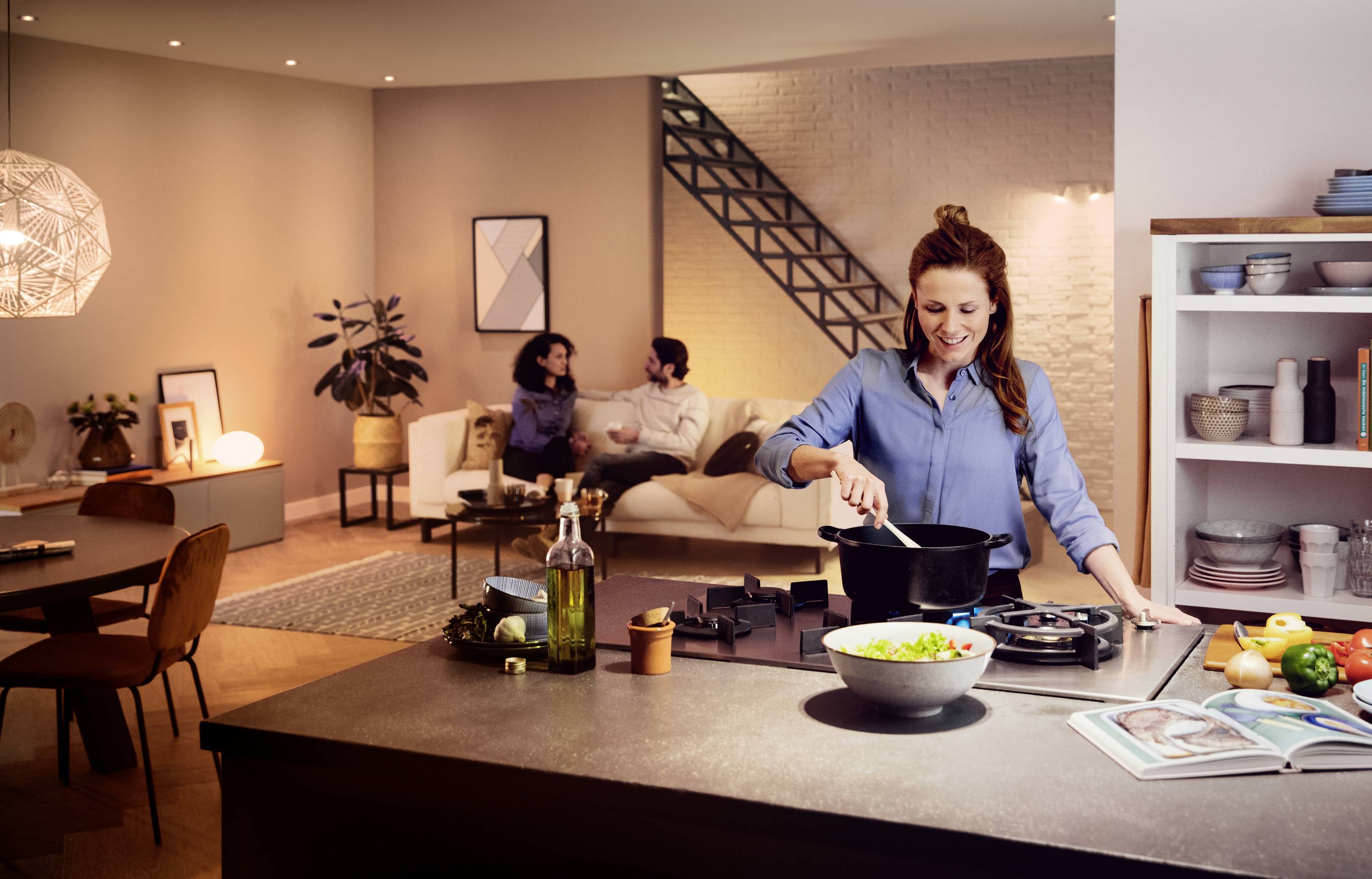 A woman is cooking in a modern kitchen, while a couple sits on a sofa in the background, chatting.