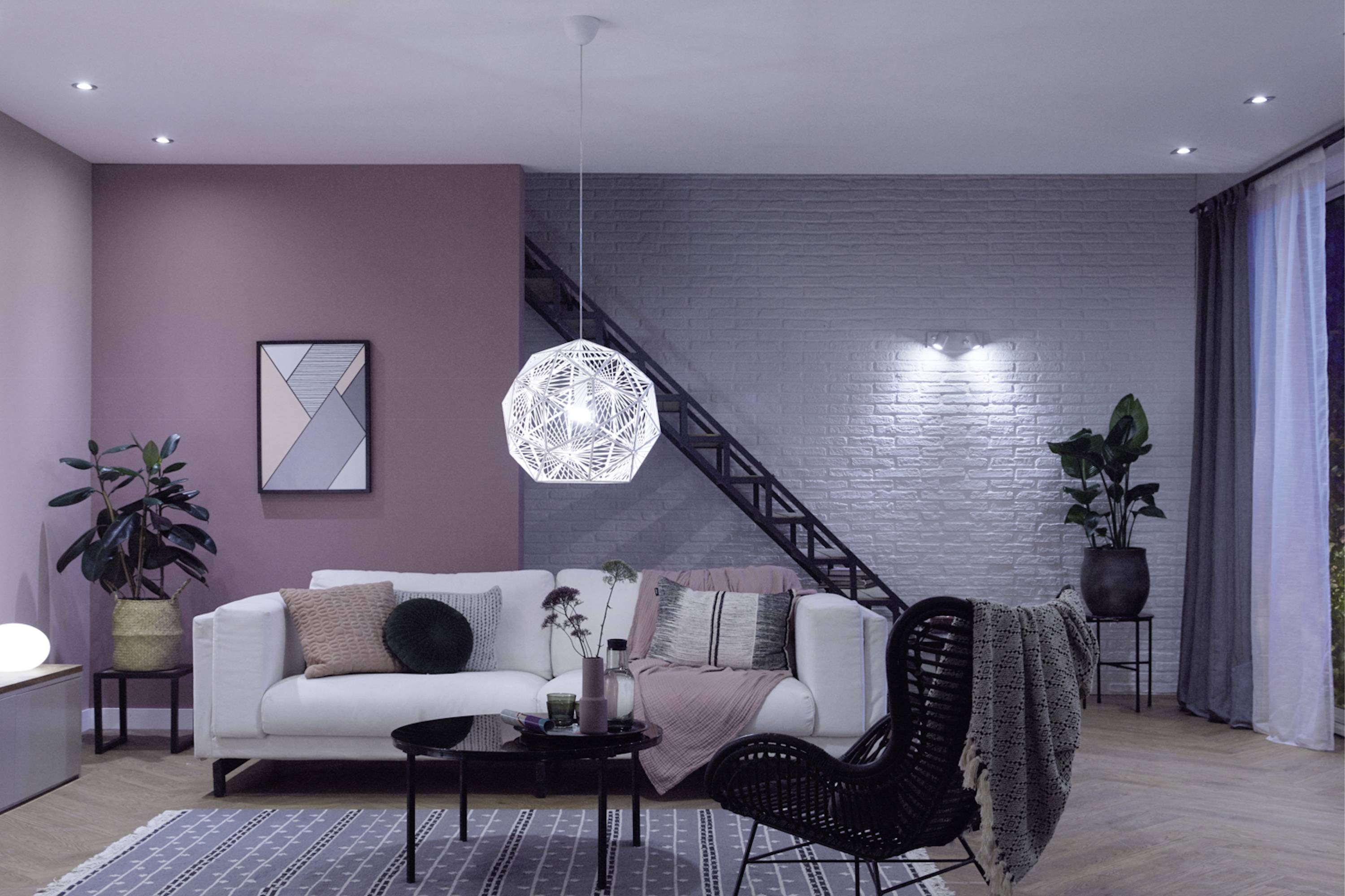 A modern living room with a white sofa, black and white lamp, black wicker armchair and plants. Pink and grey walls in the background.