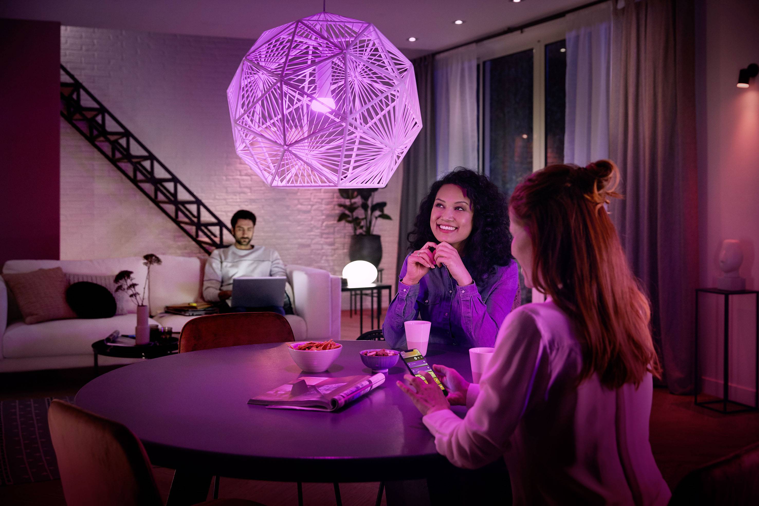 A group of three people are relaxing in a living room with modern, purple lighting. Two are chatting at the table, while one sits on the sofa in the background with a laptop.