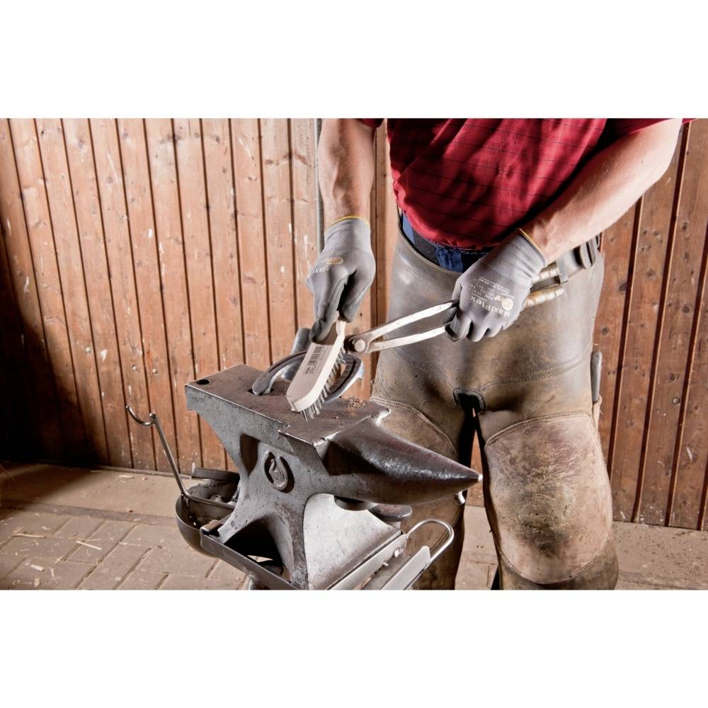 A blacksmith cleans a horseshoe with a wire brush on an anvil in a wooden workshop.