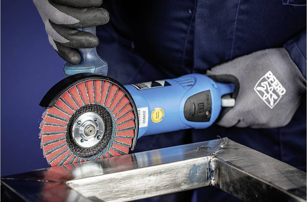 A person in work attire is grinding the edge of a metal piece with a blue angle grinder. Gloves are protecting their hands.