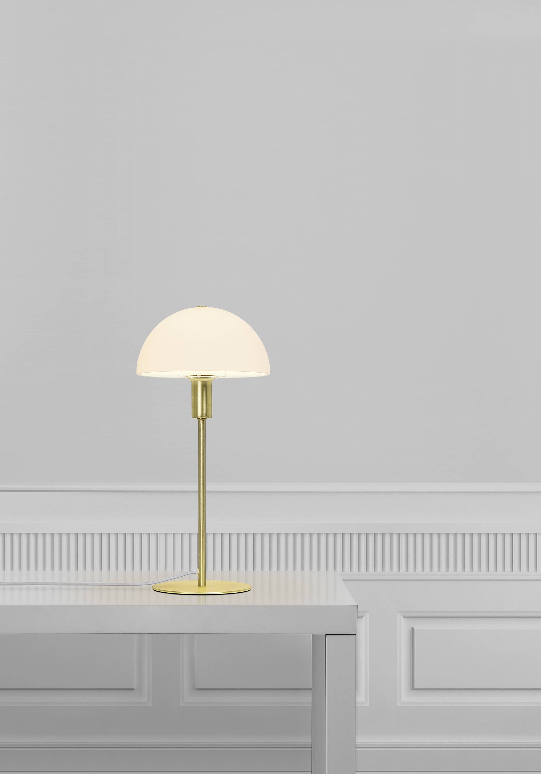 A minimalist table lamp with a golden stand and white shade sits on a plain white table in front of a grey wall.