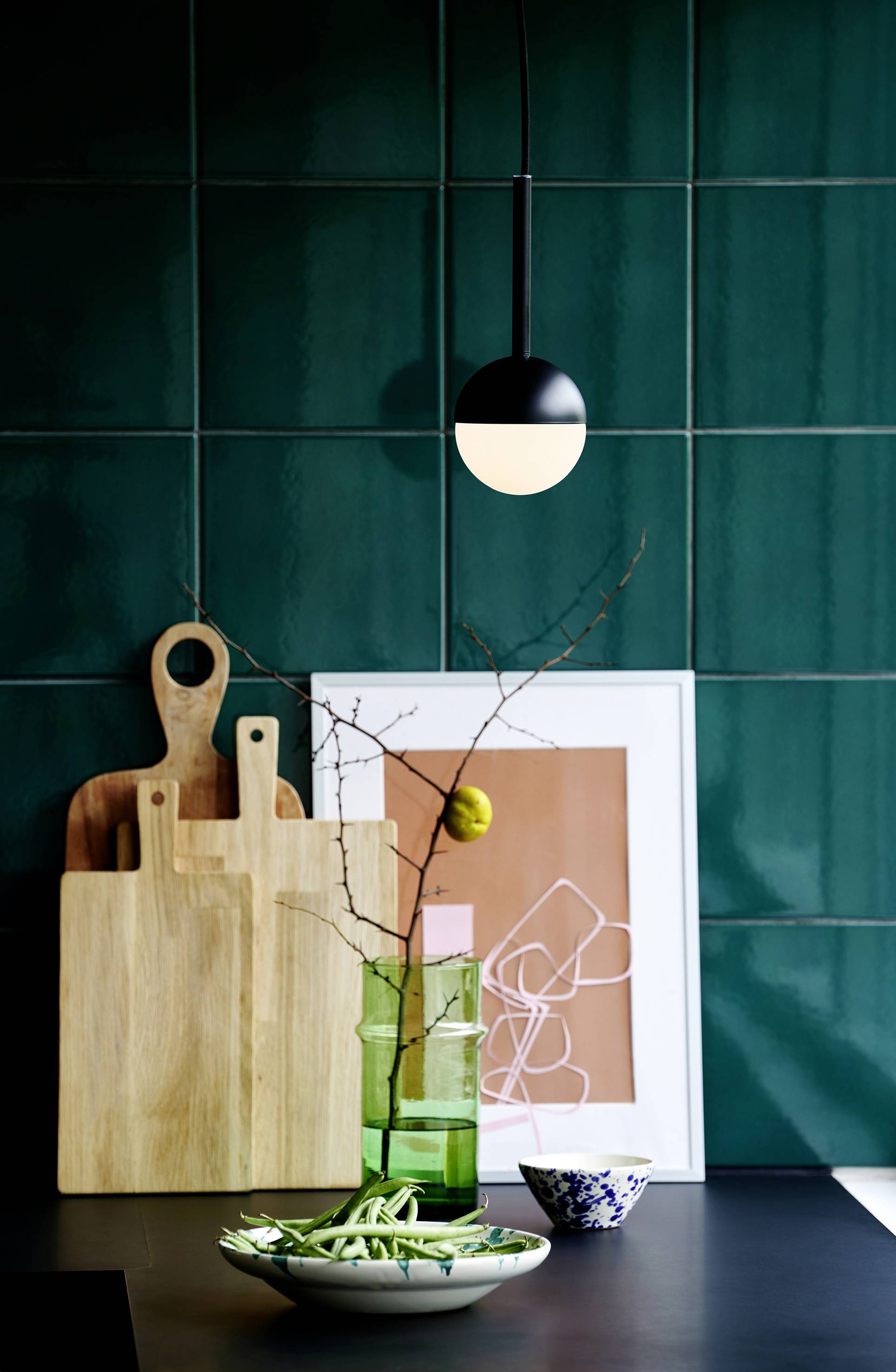 A green tiled splashback with two chopping boards, a green glass with a branch, a white bowl, and artwork in the background.