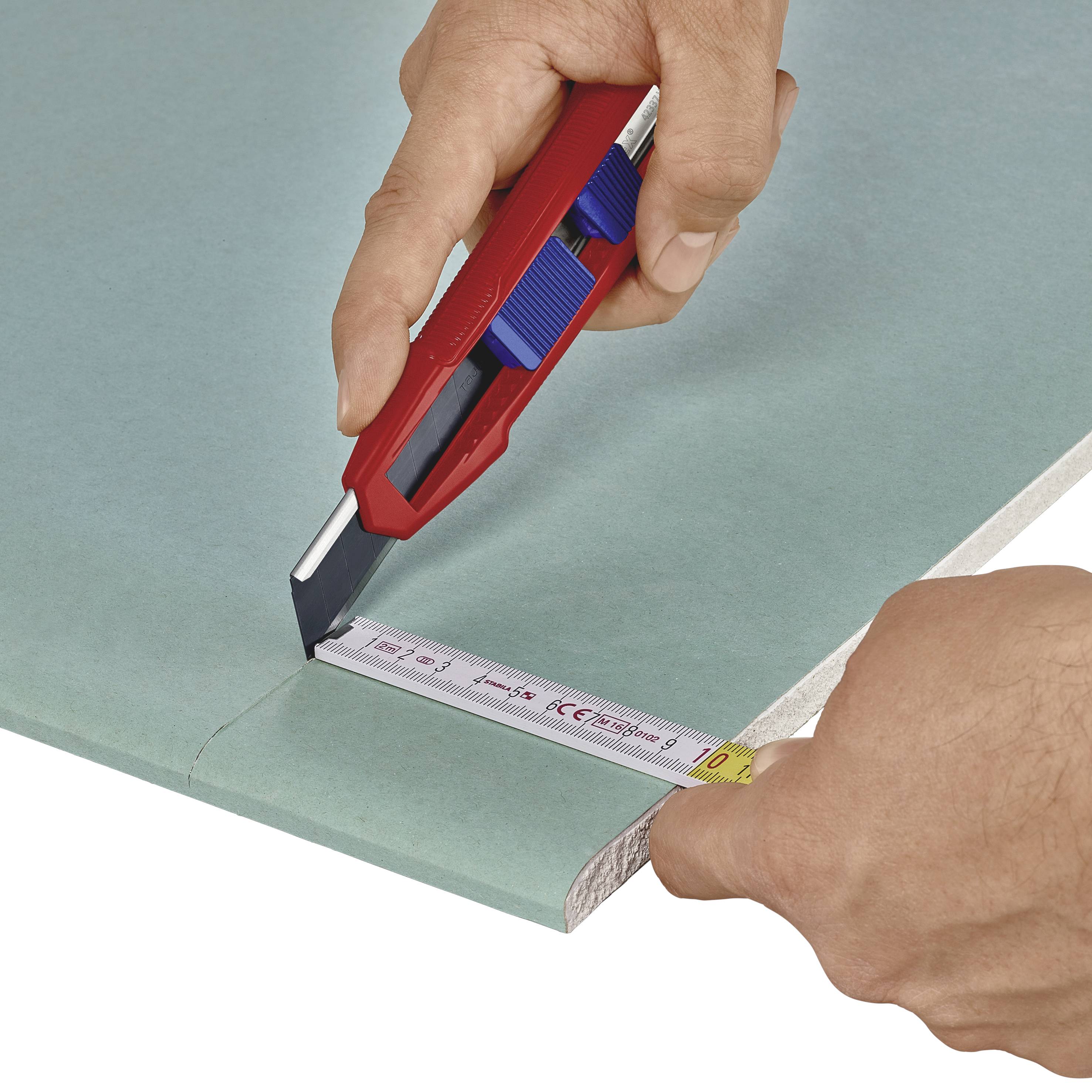 A person is cutting a piece of green plasterboard with a red utility knife along a ruler.