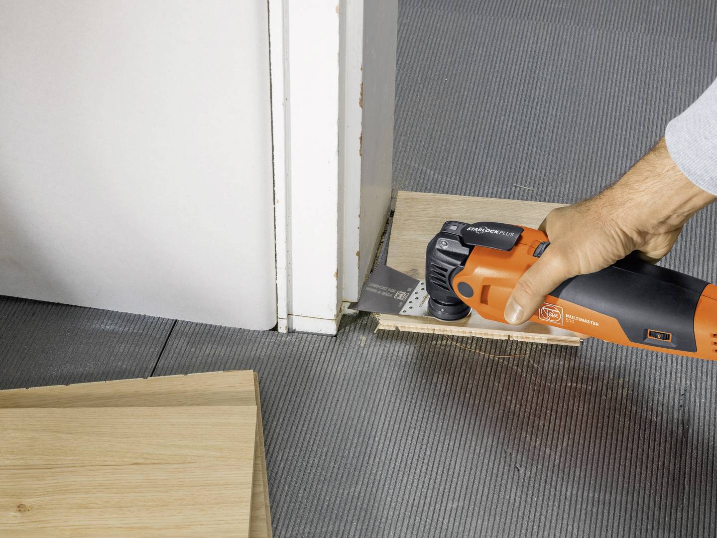 A person is using an oscillating multi-tool to work on a piece of wood whilst it lies on the ground next to a door.