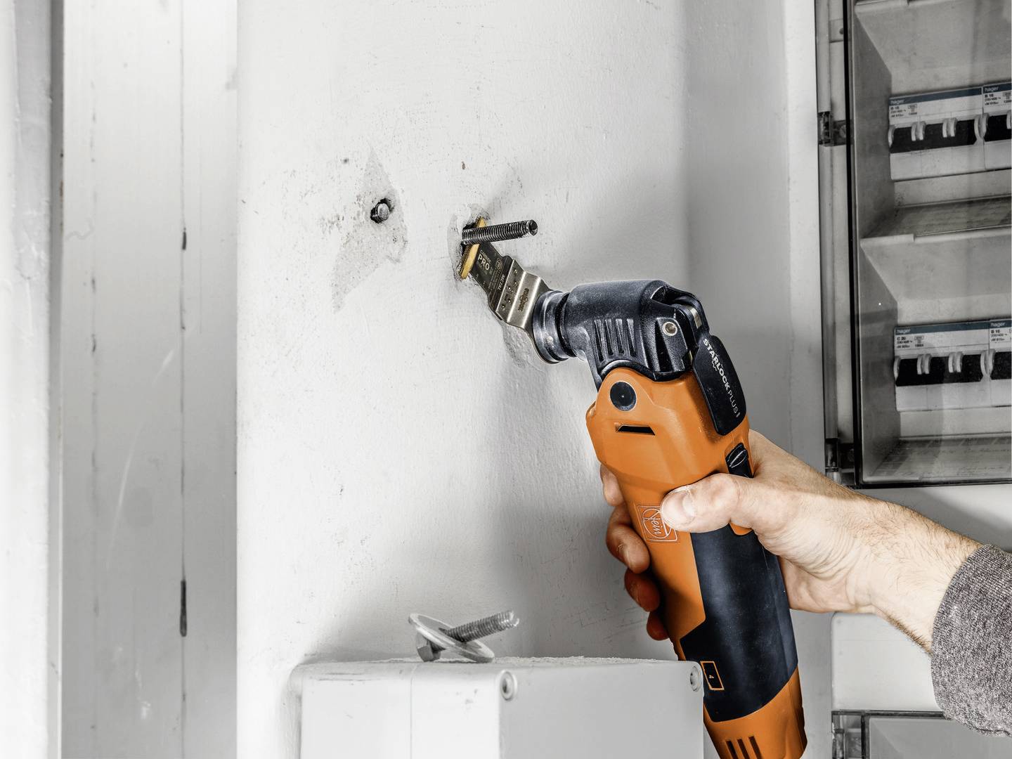 A person is using an orange multi-tool to remove a screw from a white wall.