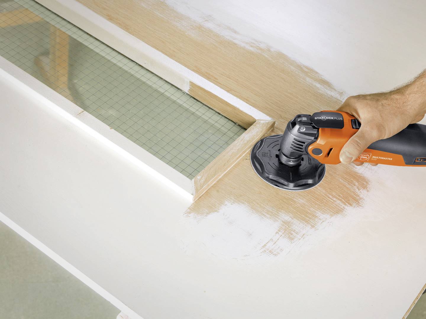 A right hand is using an orange electric tool to sand smooth white surfaces on wood. A measuring tape is positioned to the left.