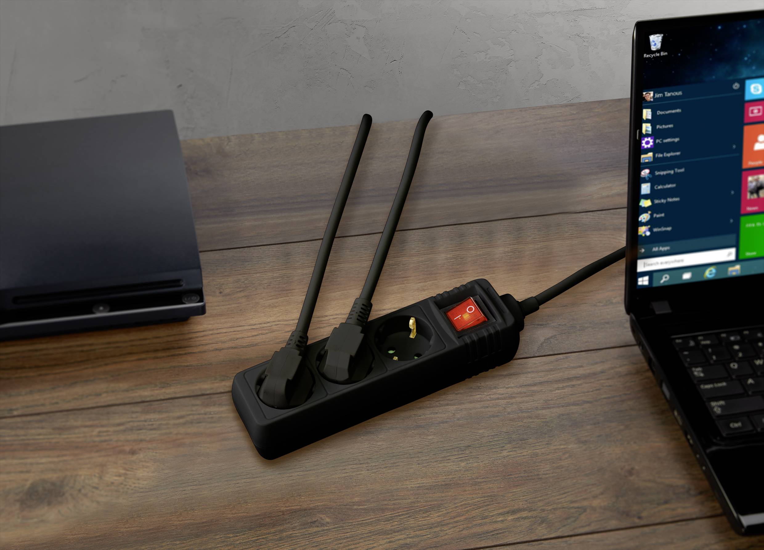 Laptop on wooden table, connected to a multi-socket extension lead. Two plugs inserted, one socket free. Closed device on the left, possibly another laptop.