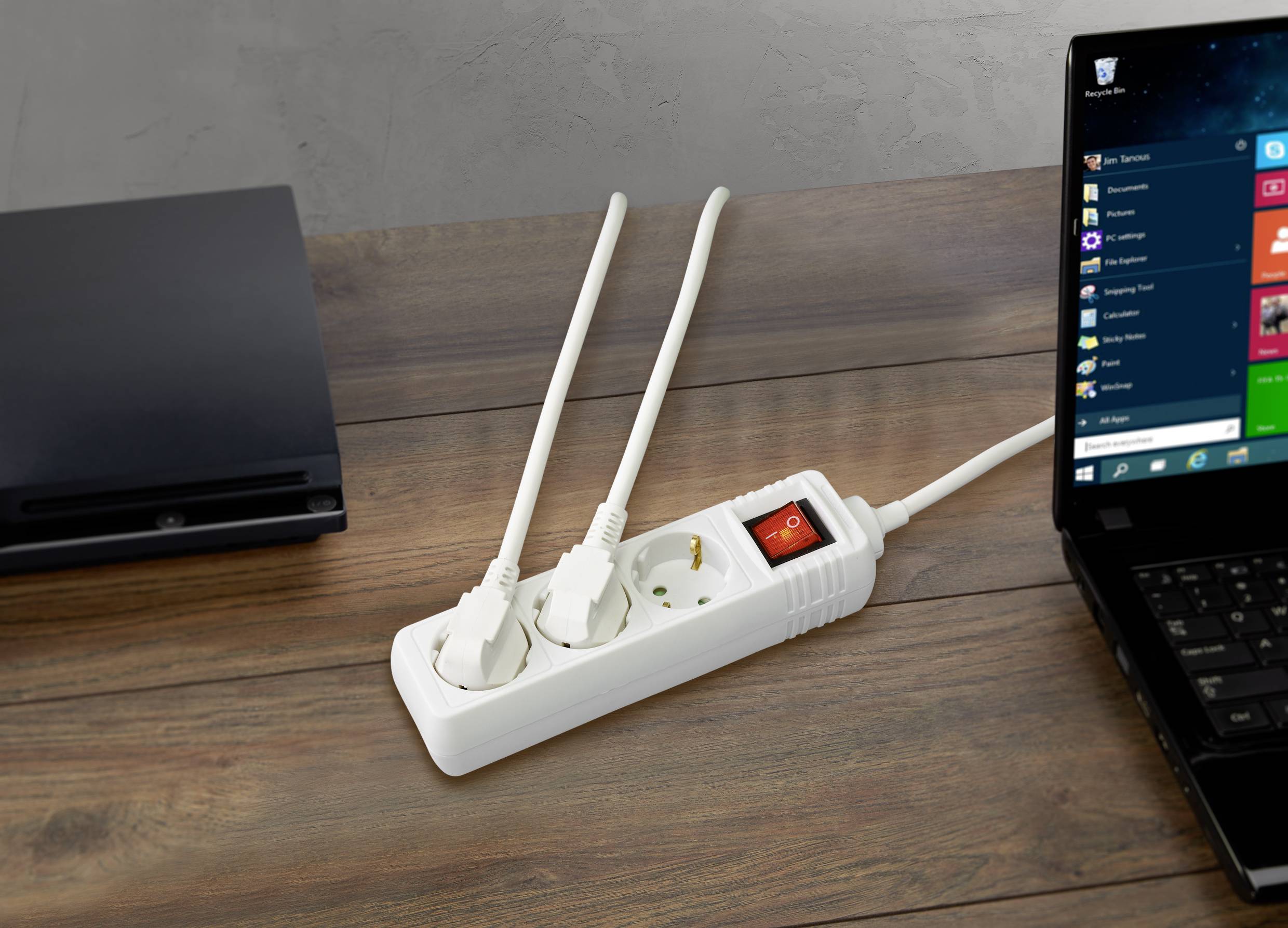 'Multi-socket extension lead with two plugged-in cables next to a laptop on a wooden table. The socket's switch is set to 'On'.'