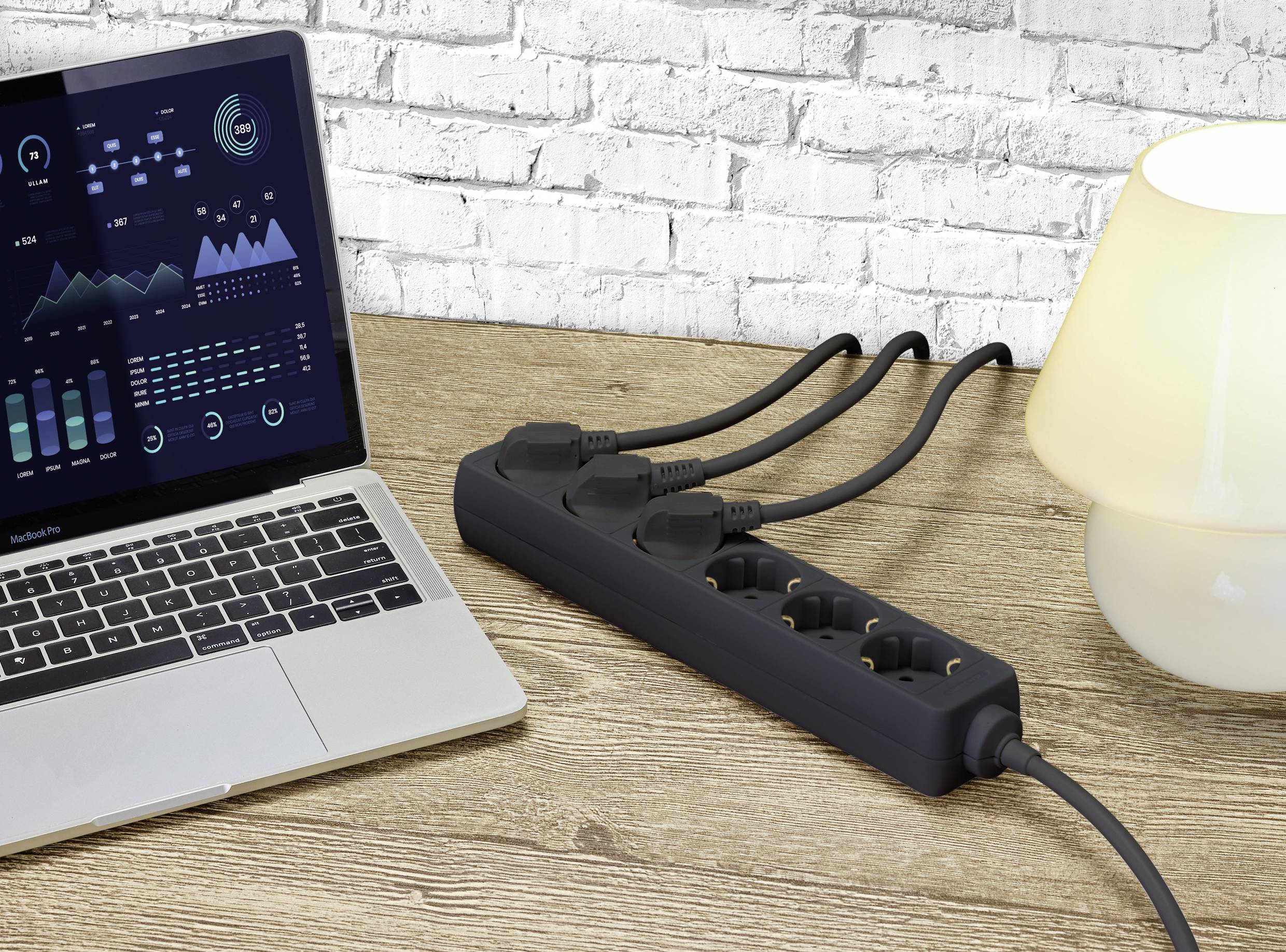 A laptop with data display sits on a wooden table next to an illuminated lamp and a black power strip.