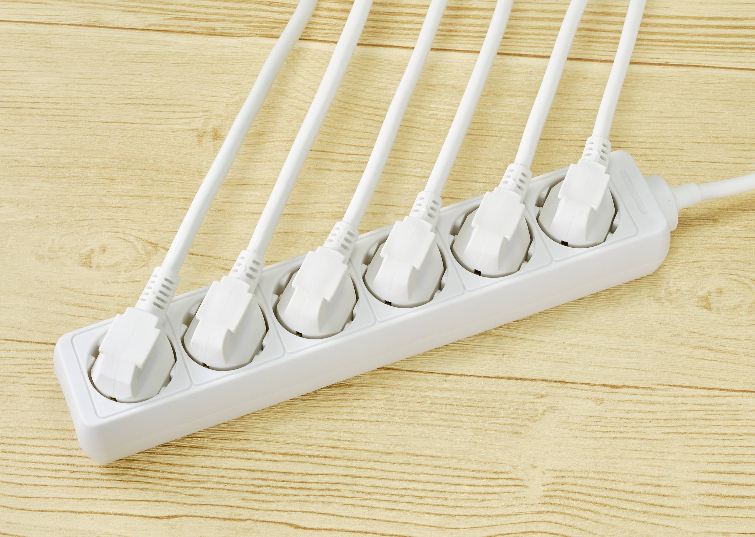 A white extension lead with six occupied sockets lies on a wooden floor. All sockets are fitted with power plugs.