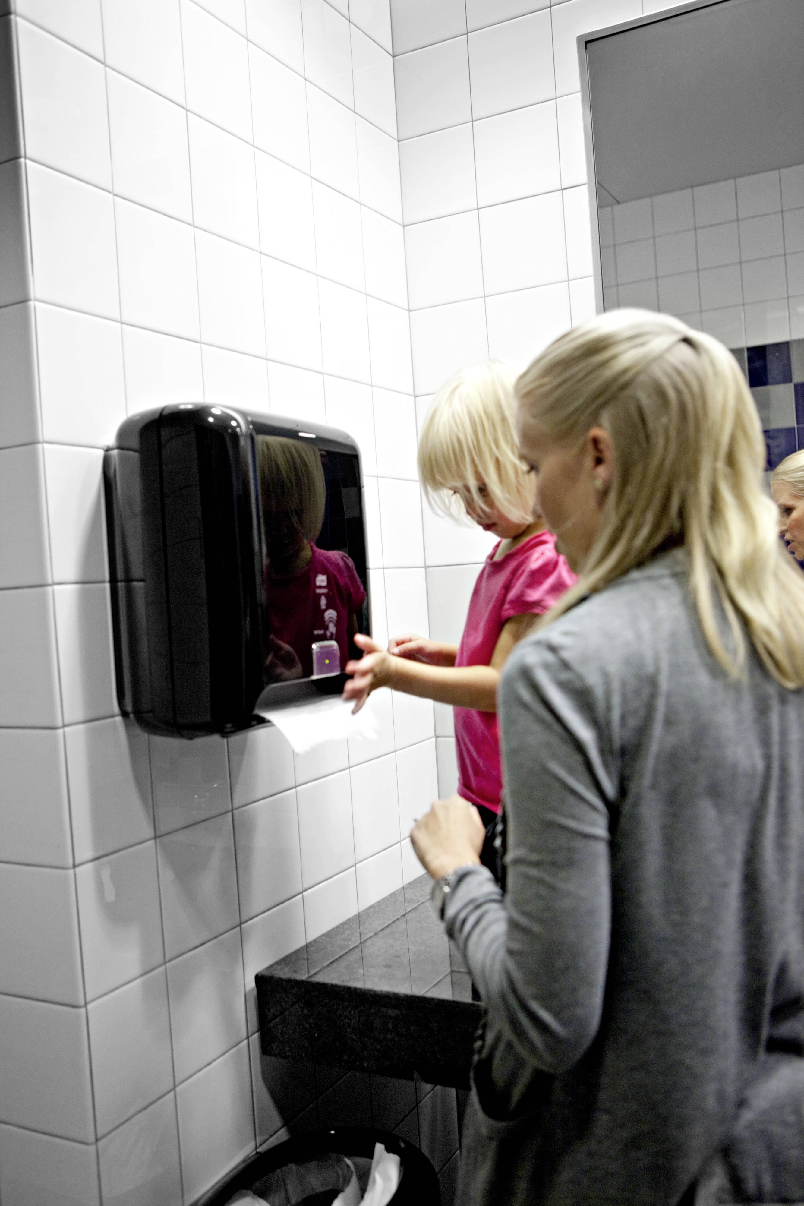 A woman is helping a small child use a hand dryer in a bathroom. Both are standing in front of a tiled wall.