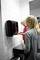 A woman is helping a small child use a hand dryer in a bathroom. Both are standing in front of a tiled wall.