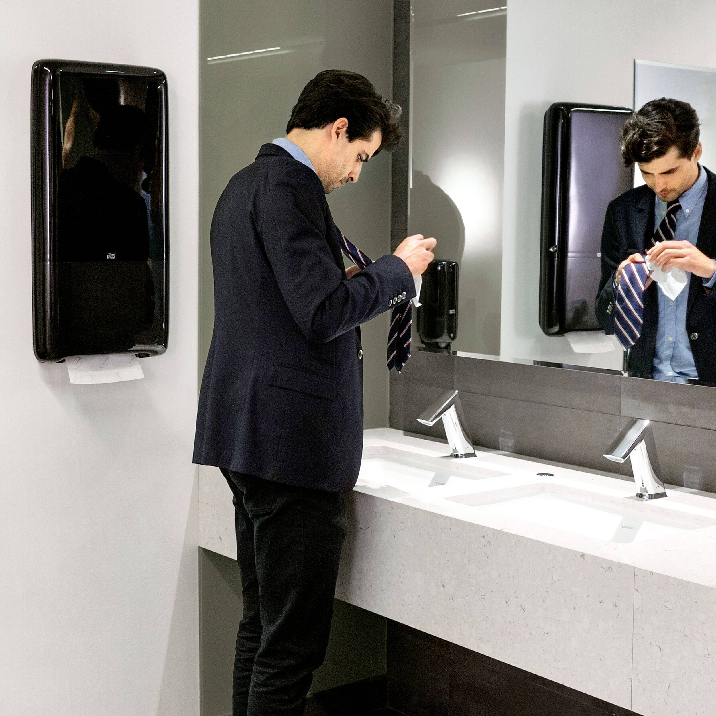 A man in a suit stands in a bathroom in front of a mirror, tying his tie. A towel dispenser is mounted on the wall.