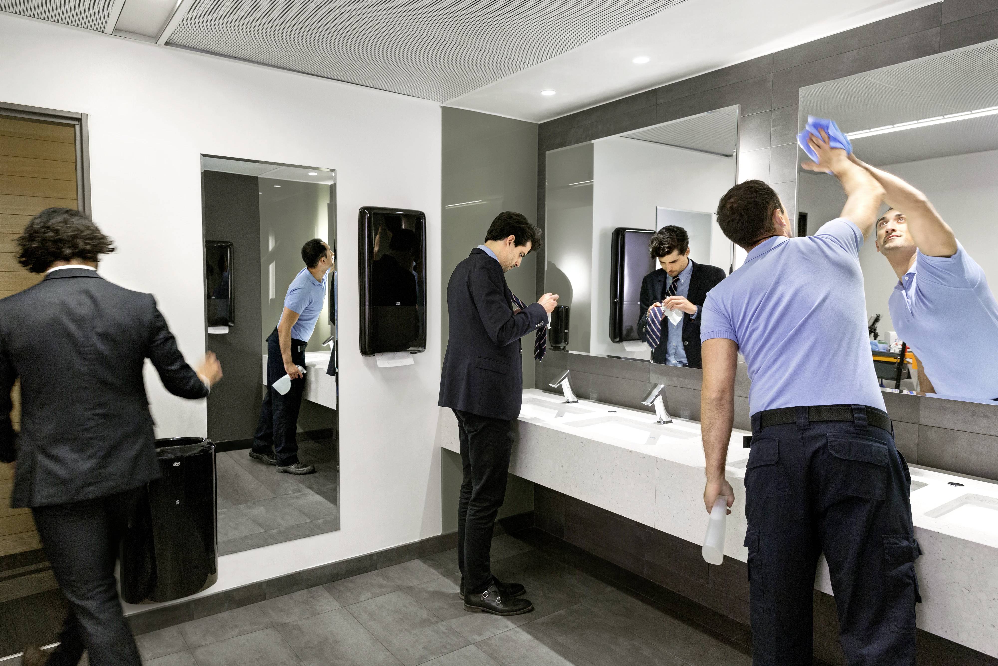 Several people in suits or uniforms are in a public lavatory. One person is cleaning the mirror, while others are washing their hands.