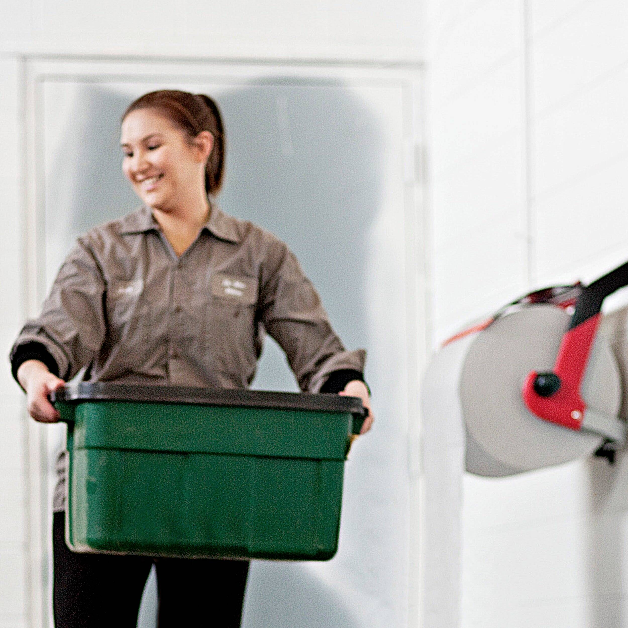 A person is carrying a green container in an industrial setting. White walls and machinery are visible in the background.