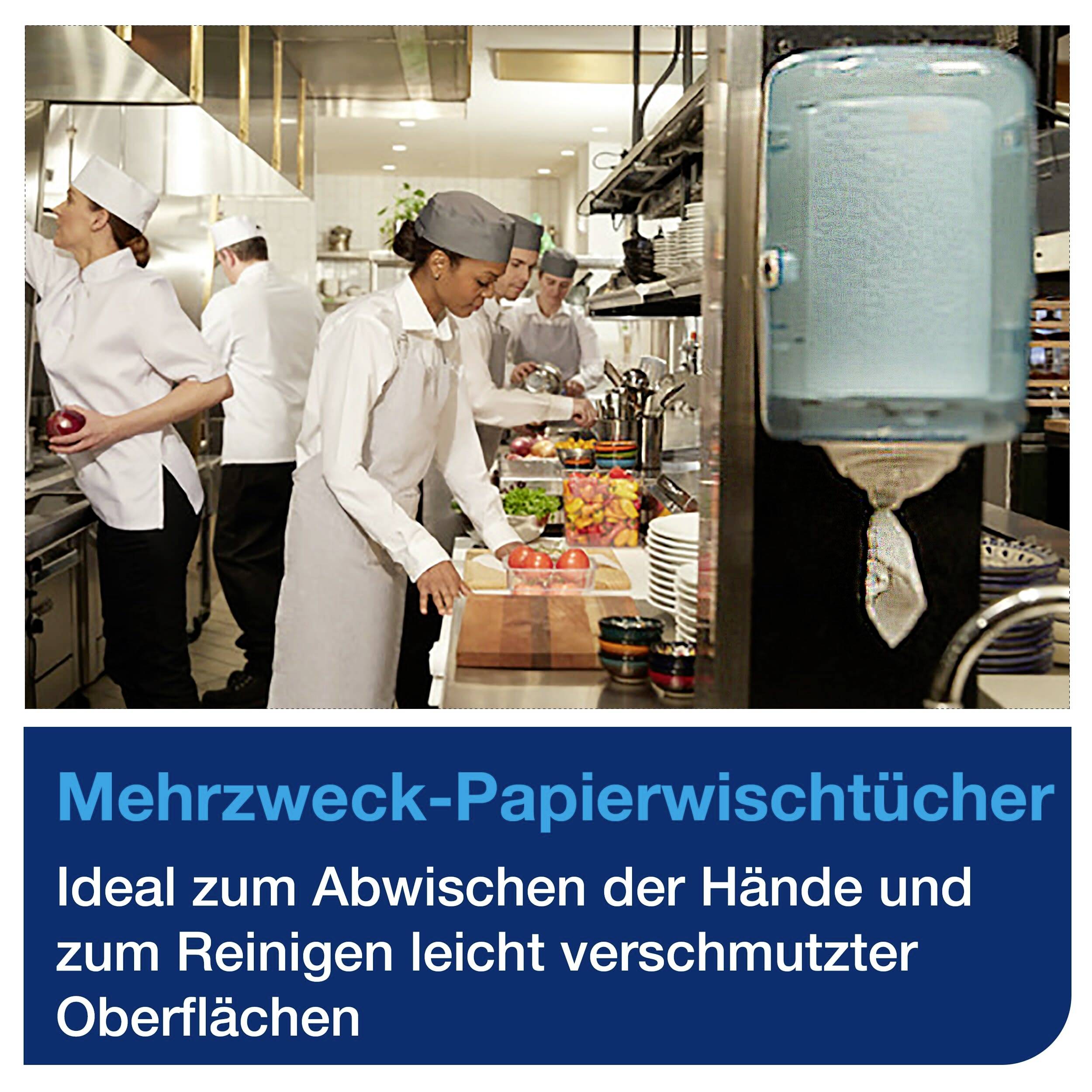 Chefs in a restaurant kitchen are preparing food. To the right, a dispenser for multi-purpose paper towels with a label underneath.