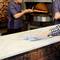 Two people in a bakery. One person is baking bread in the oven, whilst the other is cleaning the counter with a cloth.