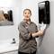 A woman in work attire pulls a paper towel from a dispenser in a public washroom. A mirror reflects her image.