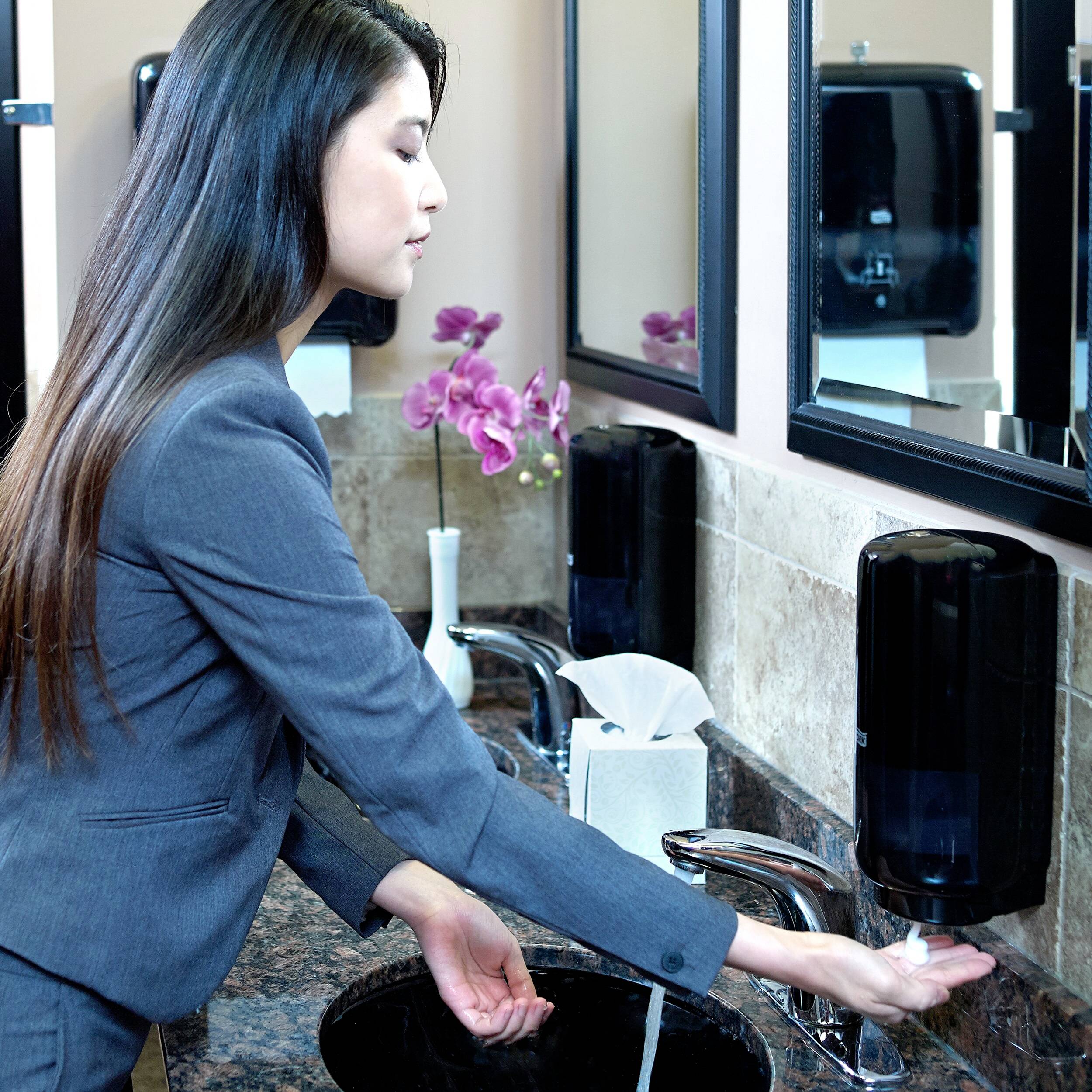 A woman in a suit is washing her hands in a bathroom. She is using a soap dispenser, and hand towel dispensers can be seen in the background.