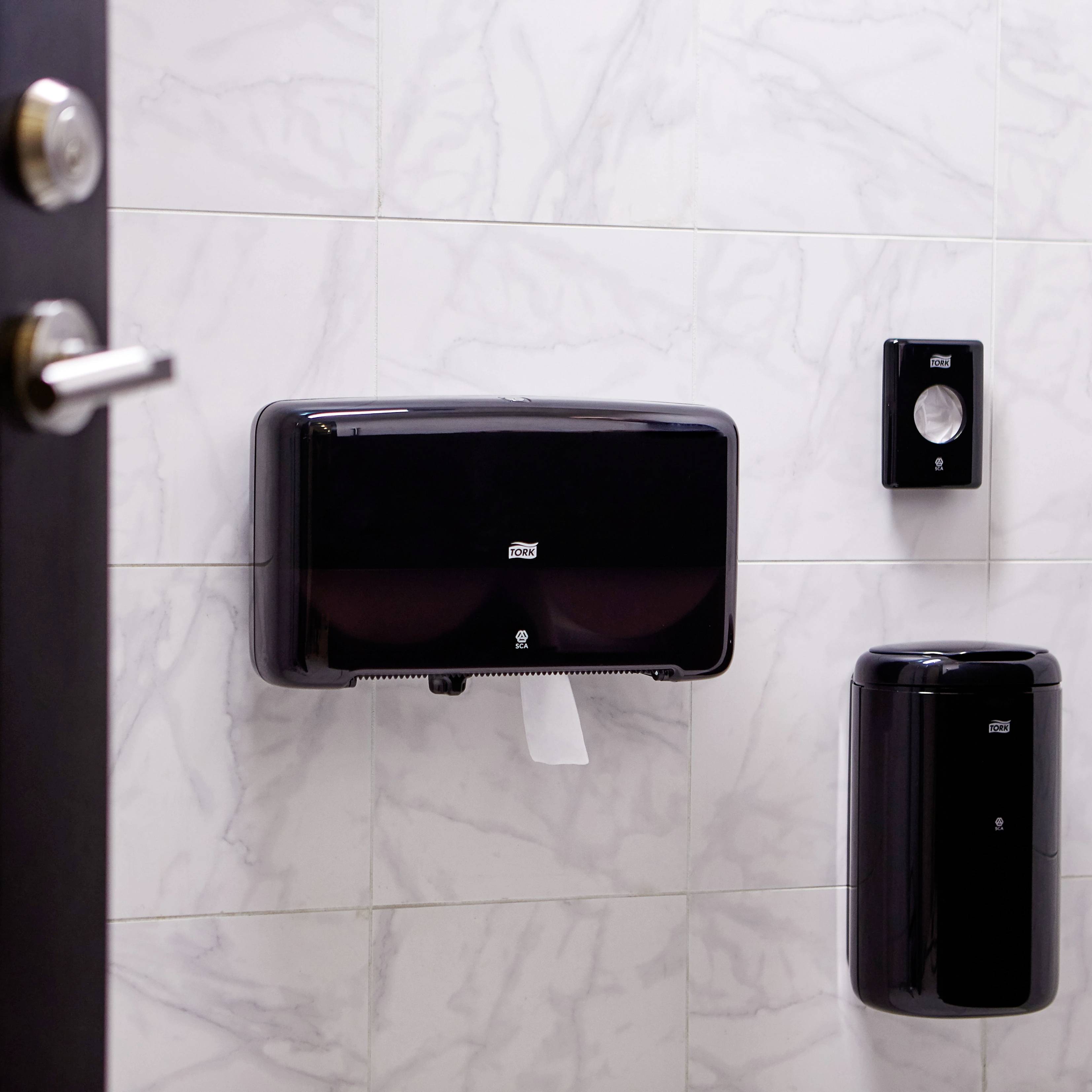 A public toilet cubicle with a paper towel dispenser, soap dispenser, and waste bin against a white tiled wall.