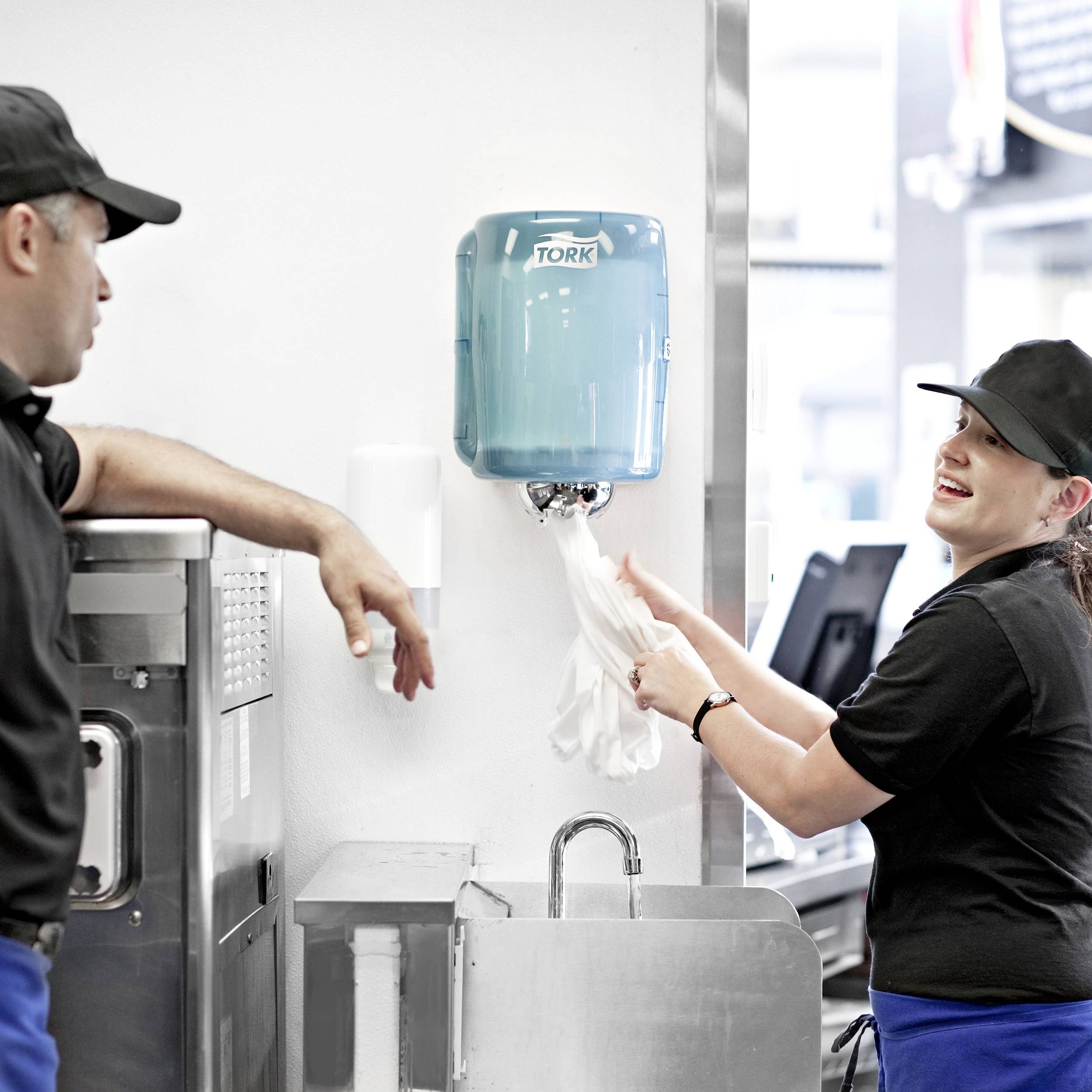 Staff in uniforms drawing paper towels from a dispenser, with the working environment suggesting a kitchen or canteen.