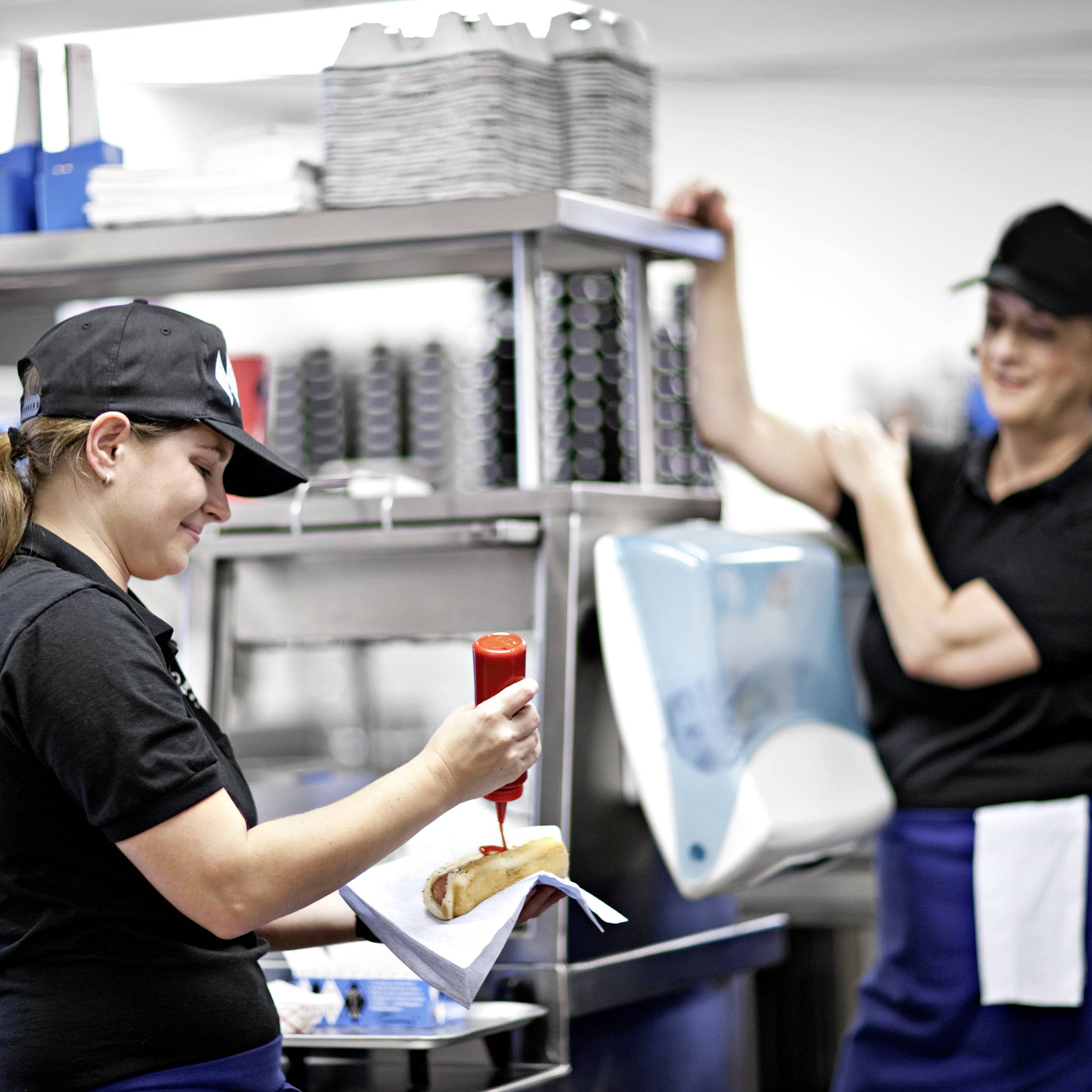 Two staff members in work attire are standing in a commercial kitchen. One person is drizzling ketchup onto a hot dog. Kitchen equipment is visible in the background.
