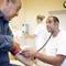 A doctor measures the blood pressure of an elderly man in a clinic. Another person in the background is operating medical equipment.