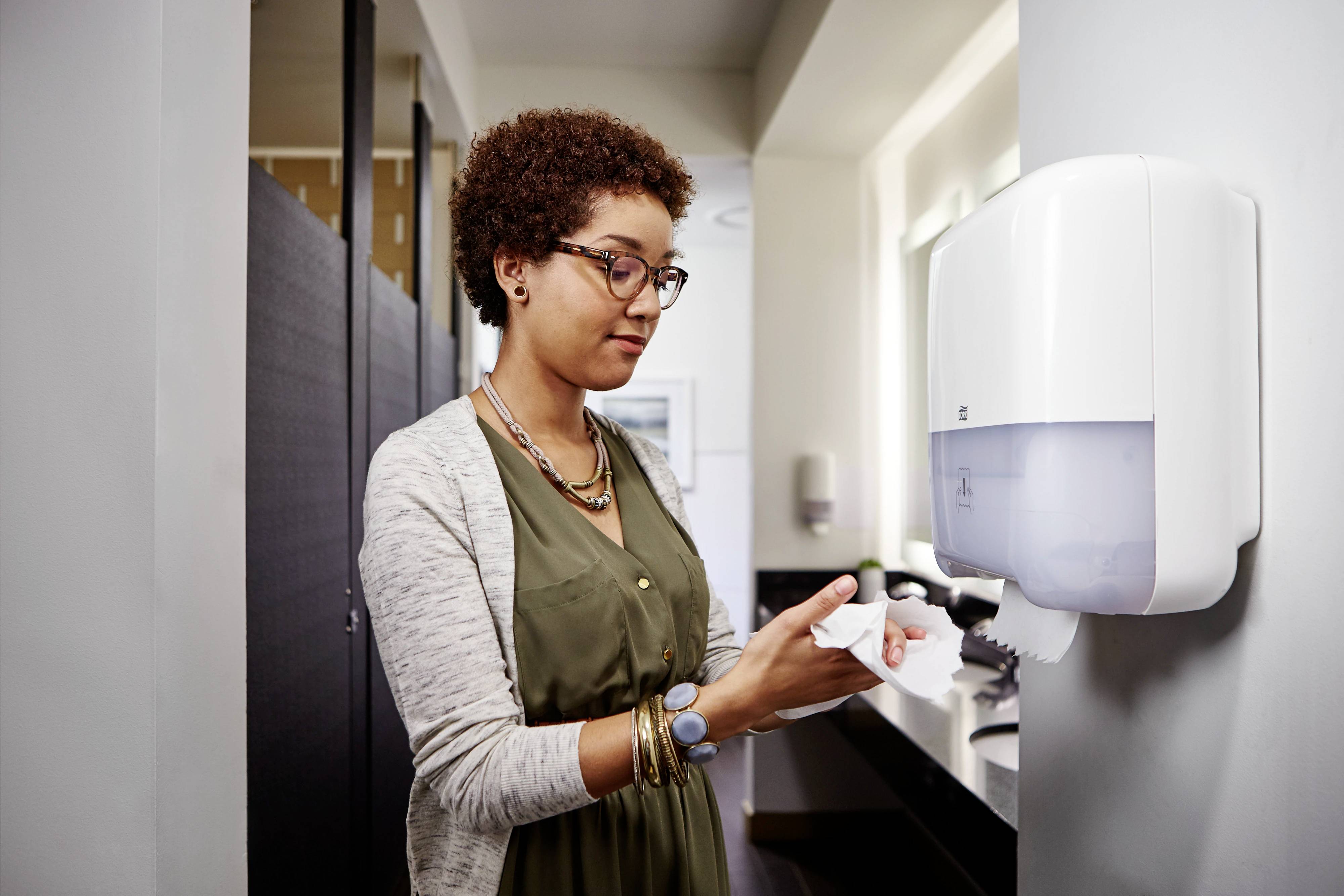 A woman is using a paper towel dispenser in a public washroom. She is wearing a green dress and a grey cardigan.