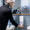 A person in a bathroom is pressing a wall-mounted soap dispenser. Blue tiles are visible in the background.