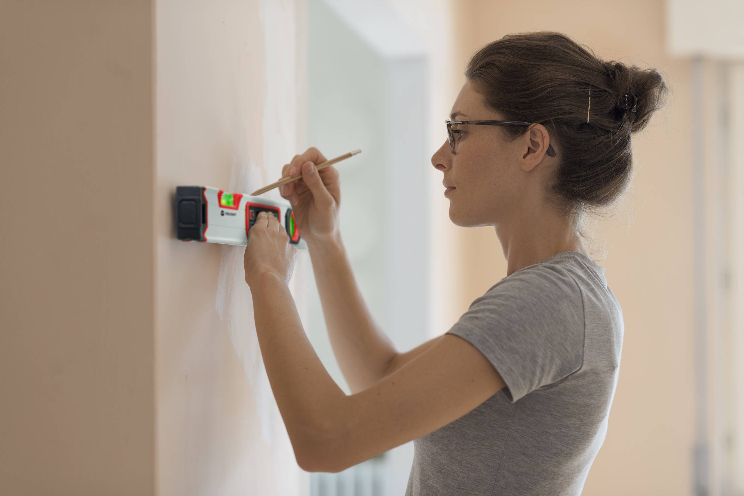 A woman is using an electronic measuring device on a wall while marking a point with a pen. She is wearing a grey t-shirt.