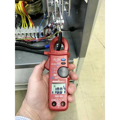 A hand is holding a red current measuring device. It displays '100.0 A' on a screen and is measuring the current of a cable in an electrical cabinet.