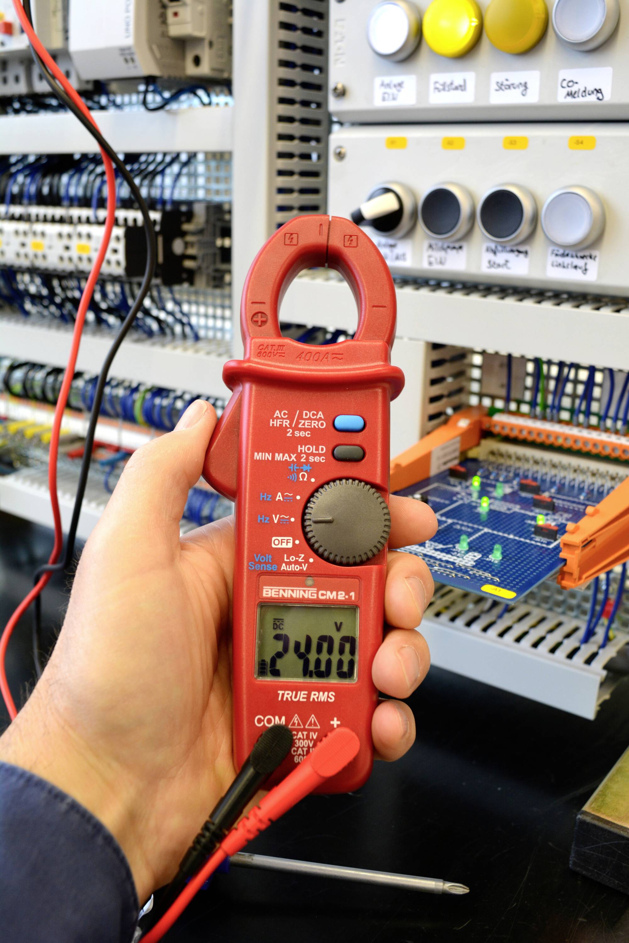 A person is holding a red digital multimeter in front of an electrical cabinet. The display shows '24.0'. The background features cables and electrical circuits.