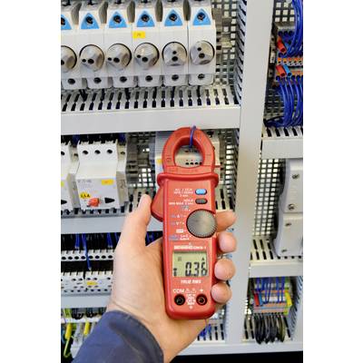 A person is measuring the electric current in a control cabinet using a red digital multimeter. The display shows '0.36'.