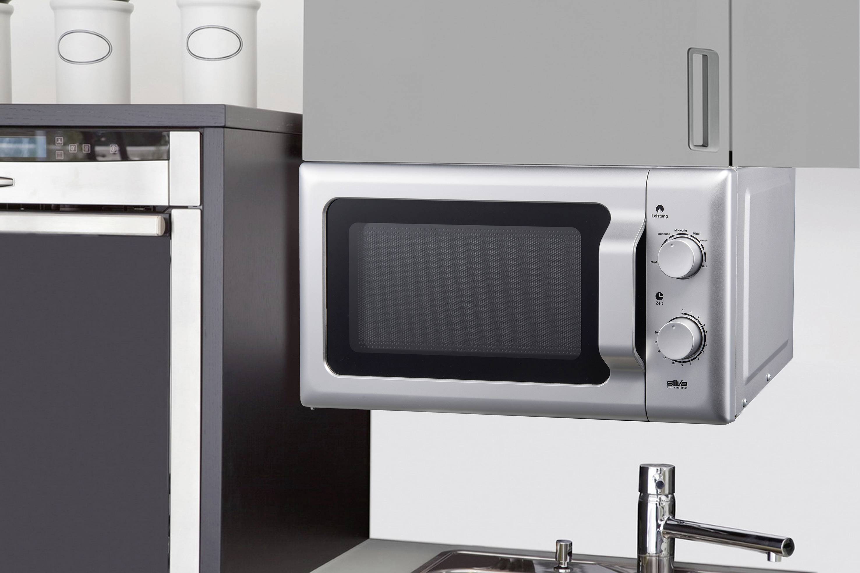 A silver microwave with two rotary knobs is mounted under a kitchen cupboard, next to a sink.