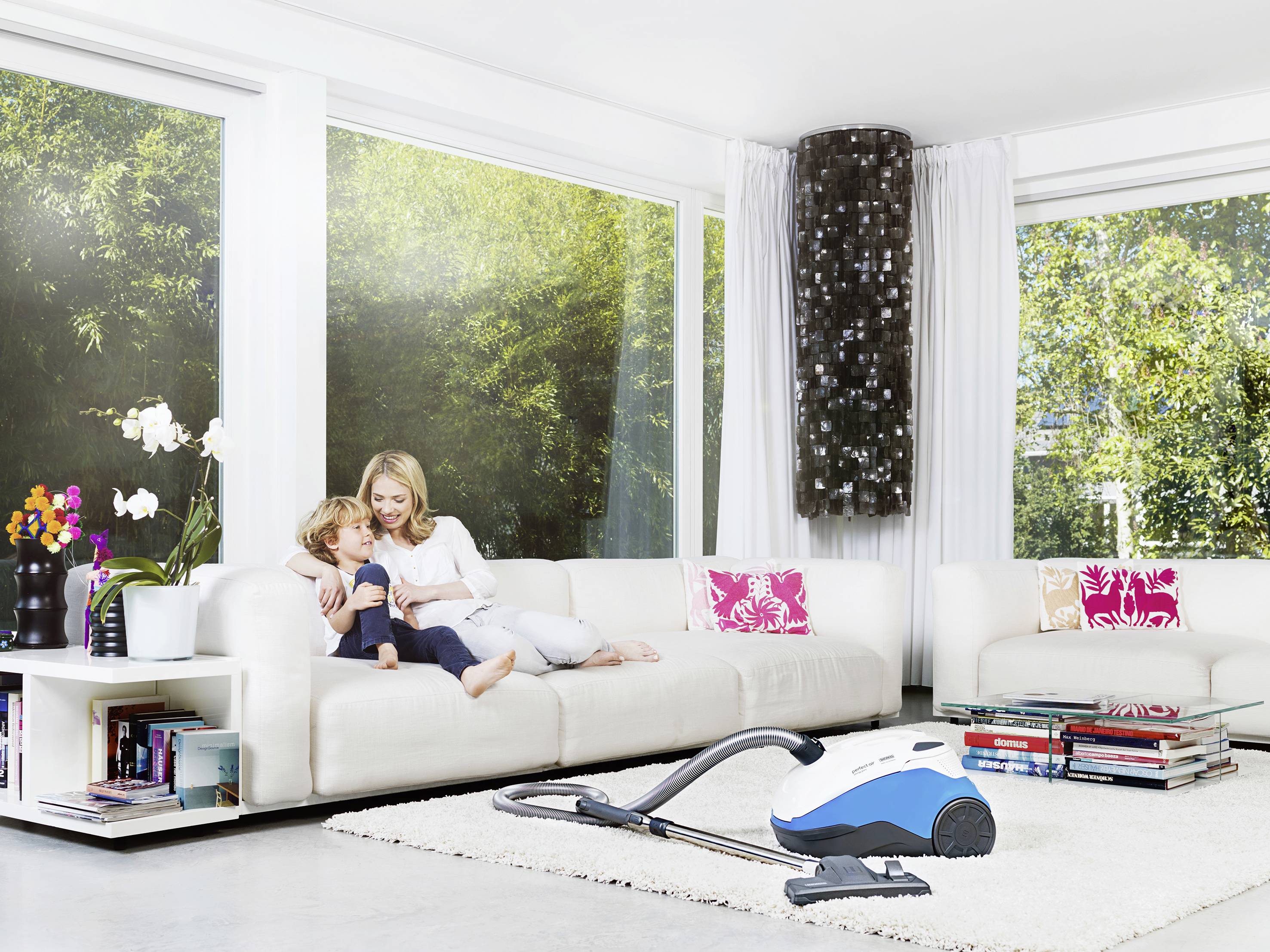 A woman is sitting with a child on a white sofa in a bright living room. A blue vacuum cleaner is standing on the floor.