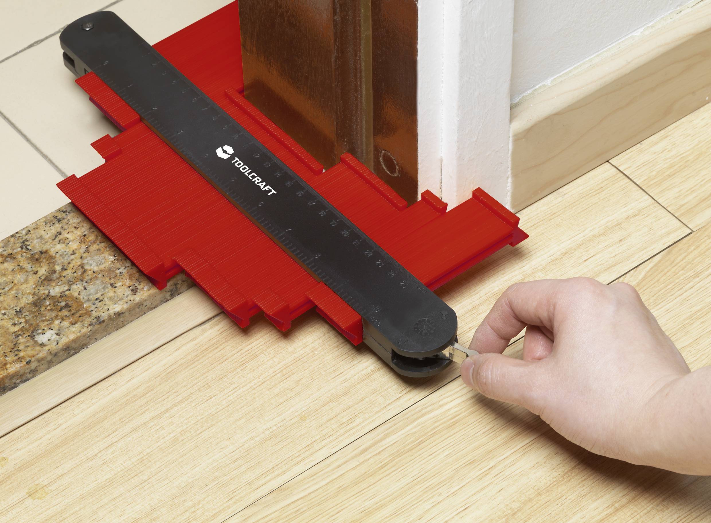 A person is using a red contour gauge tool to measure the shape of a door threshold on a light wooden floor.
