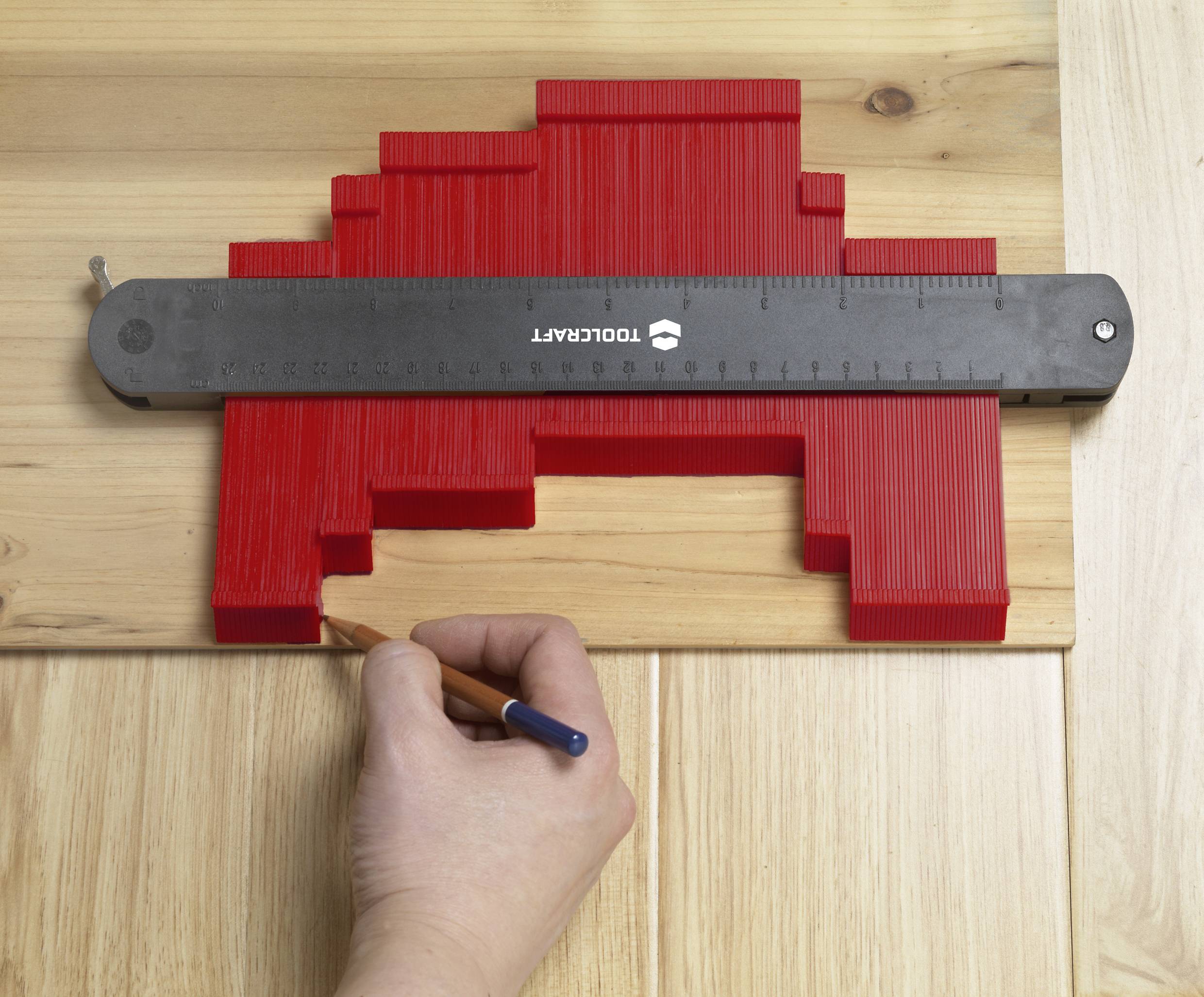 A red contour guide is lying on wood, with a hand making markings with a pencil.
