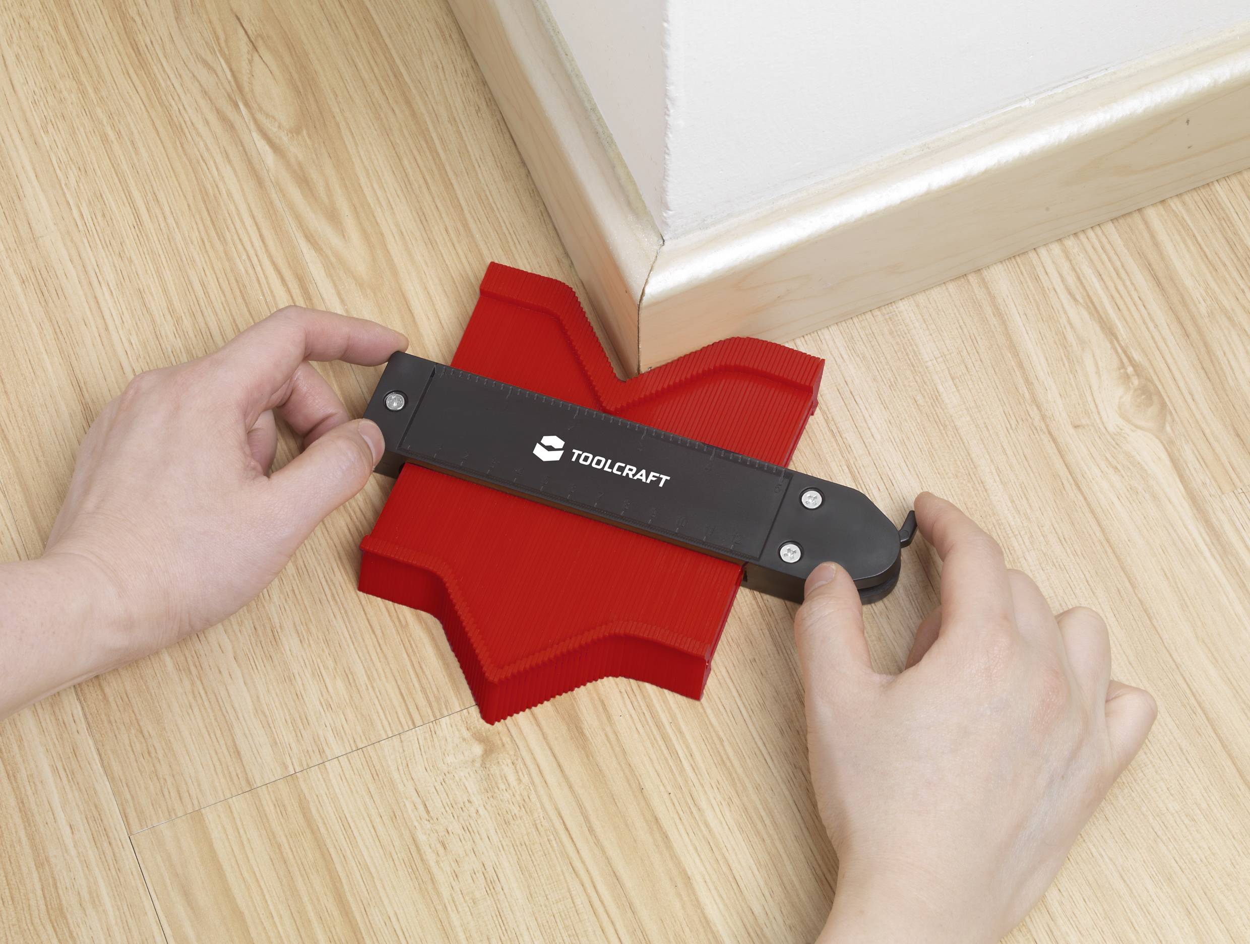 A person is using a red contour gauge tool to measure the angle shape of a wooden skirting board.
