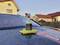 A spray bottle of car cleaner sits on a yellow cloth on the bonnet of a car covered in water droplets. Houses can be seen in the background.