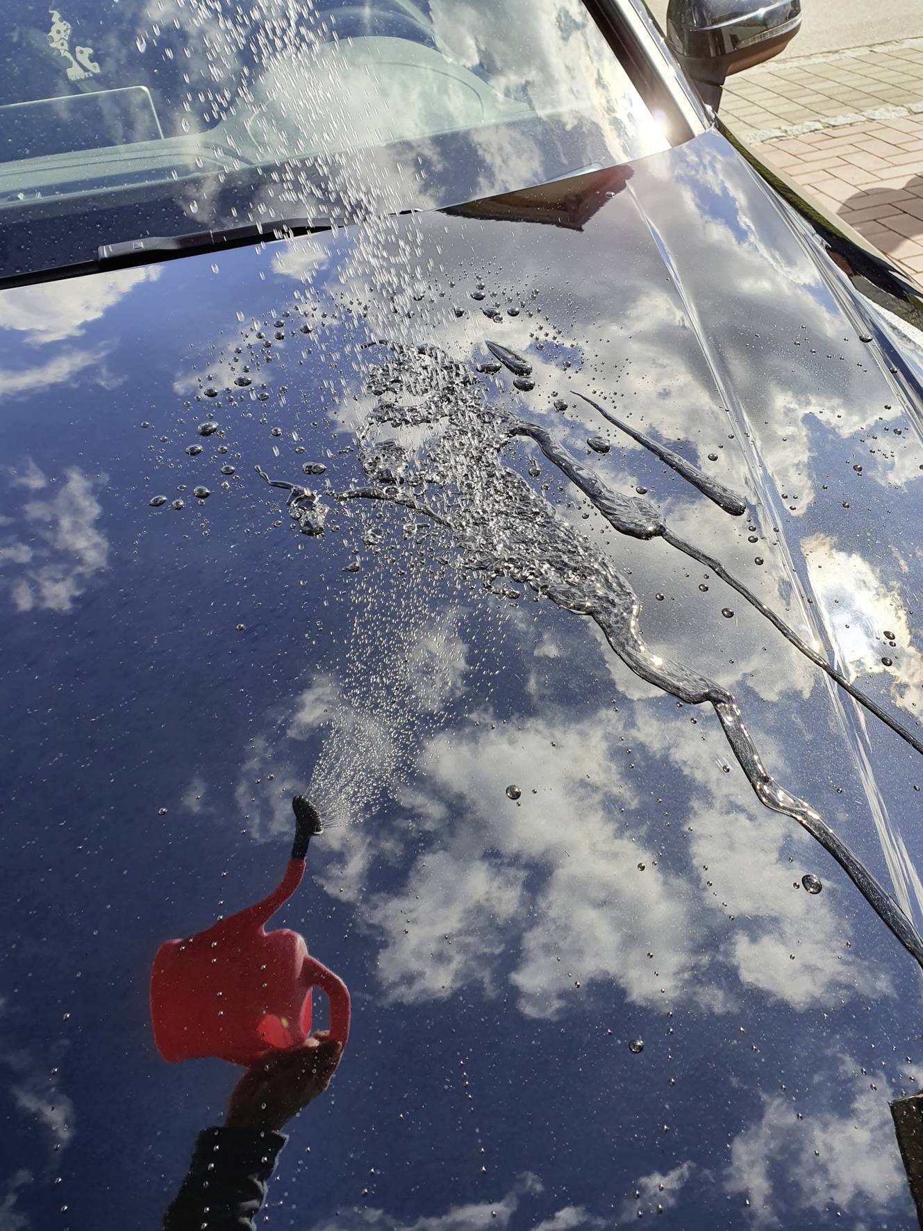 Water is being poured from a red watering can onto the gleaming bonnet of a car, with the sky and clouds reflecting on its surface.