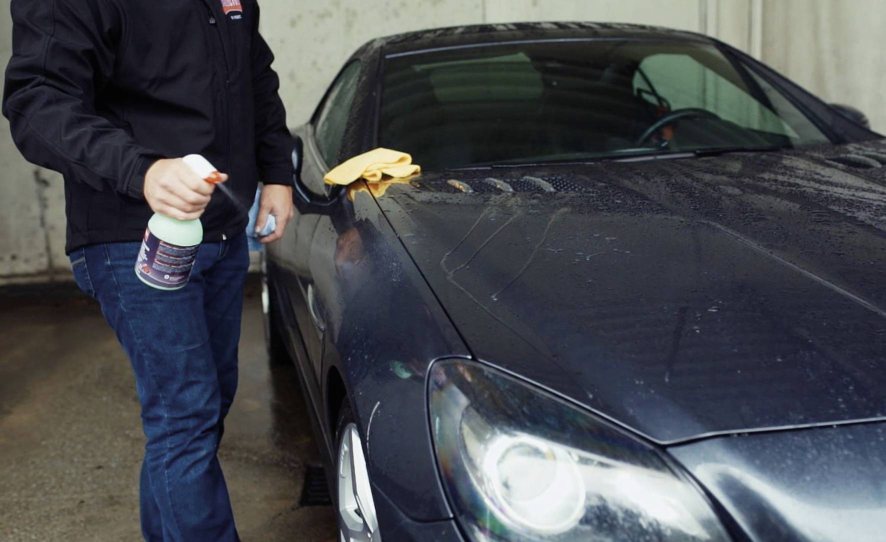 A person is cleaning the bonnet of a black car with a spray and a cloth in a garage.