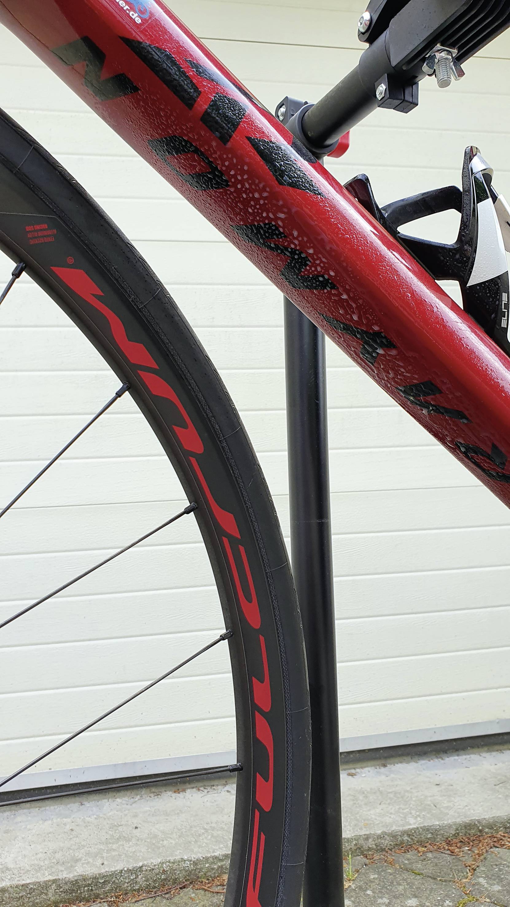 A red bicycle with black accents stands in front of a white wall. The front wheel and part of the frame are visible.
