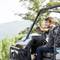 A man is sitting smiling on a quad bike in a natural setting, surrounded by trees and hills. He is holding a camera in his hand.