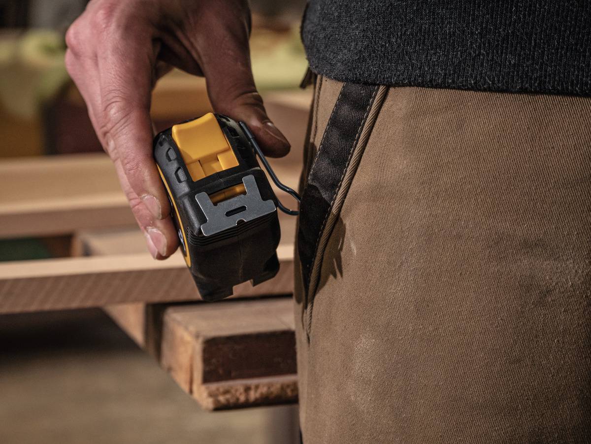 A person is holding a tape measure. They are measuring a wooden board in a workshop or woodworking environment.