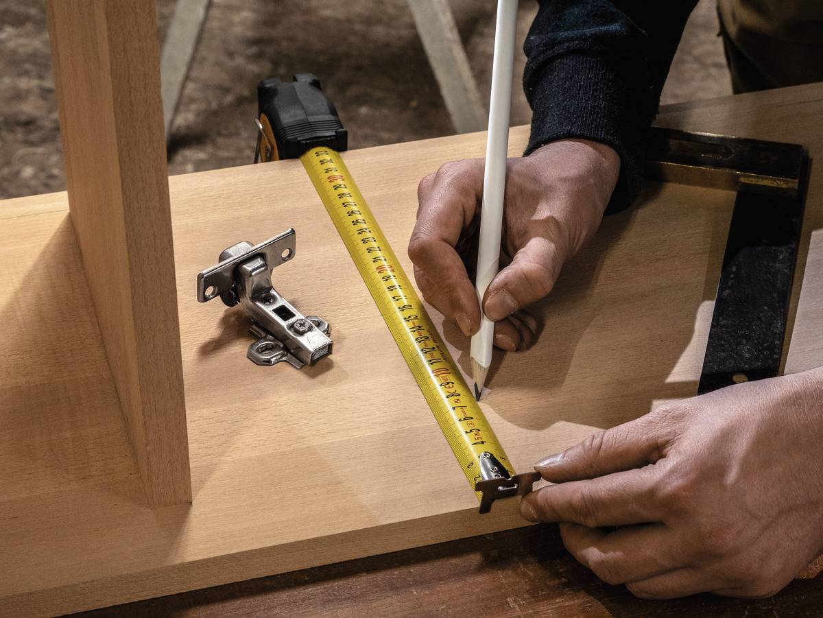 A person is measuring a wooden board with a tape measure and noting down measurements with a pencil, with a hinge lying nearby.