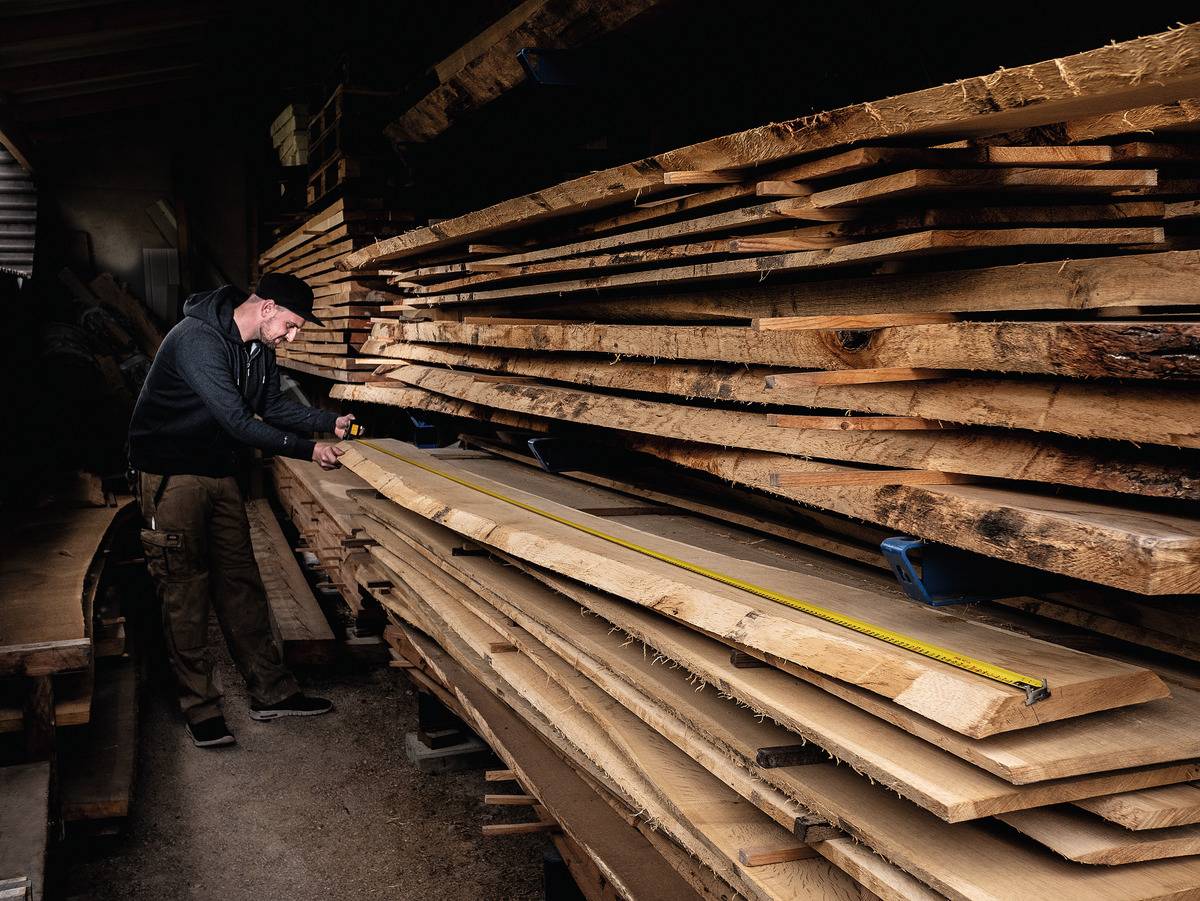 A person is measuring a long plank with a tape measure in a woodworking workshop. Stacked wooden boards can be seen in the background.