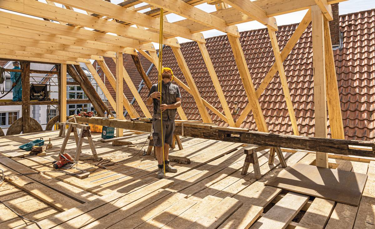 A person is working on a building site constructing a wooden house roof, surrounded by wooden beams and equipment. Roof tiles are visible in the background.