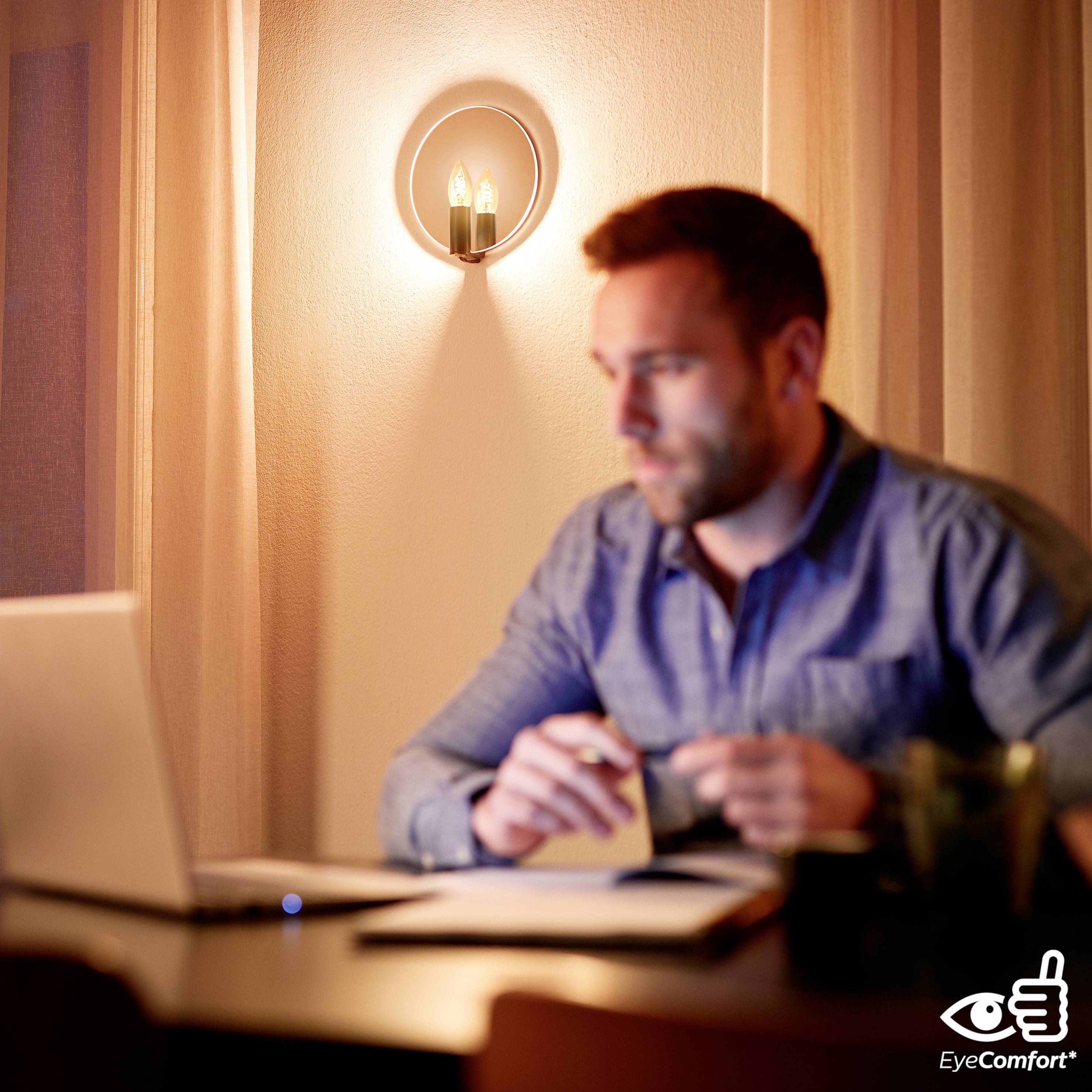 A man sits at a table in front of a laptop, holding a pen and notepad. A warm wall light illuminates the background.